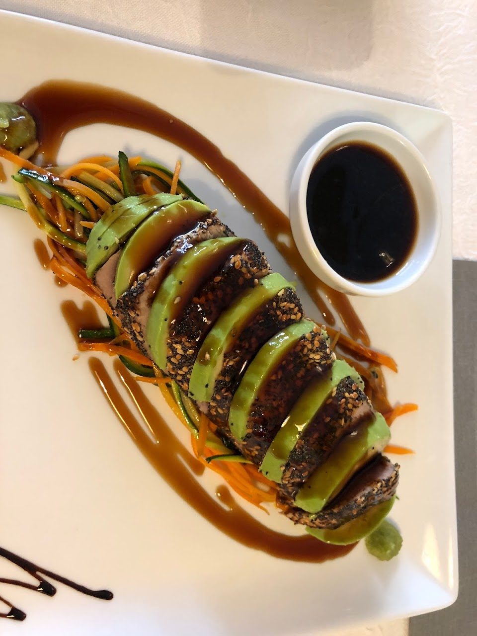 Un plat de thon en croûte de graines, tranché sur de l'avocat, avec une sauce soja et des légumes en julienne