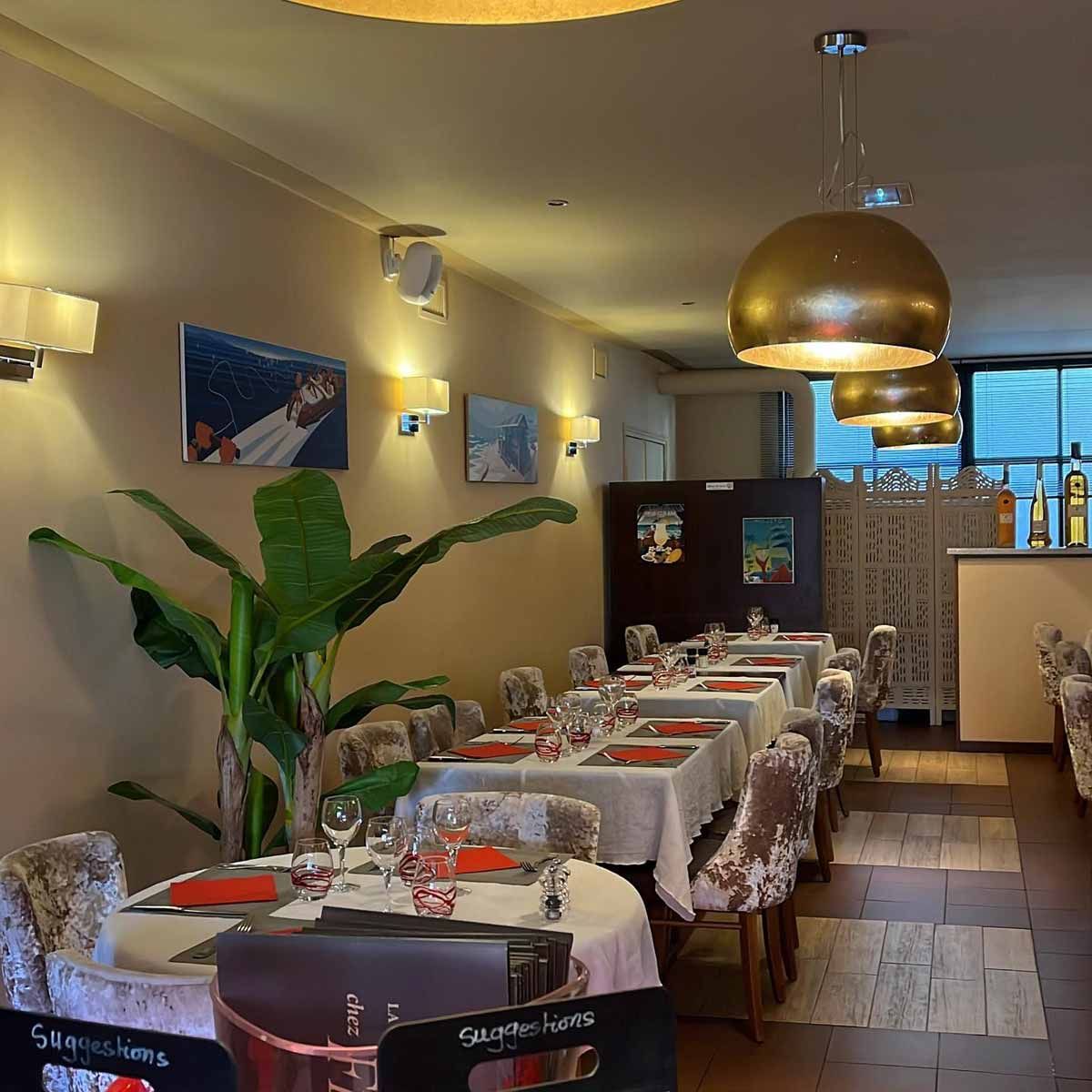 La salle : tables nappées de blanc, serviettes rouges, lampes suspendues dorées et plante verte